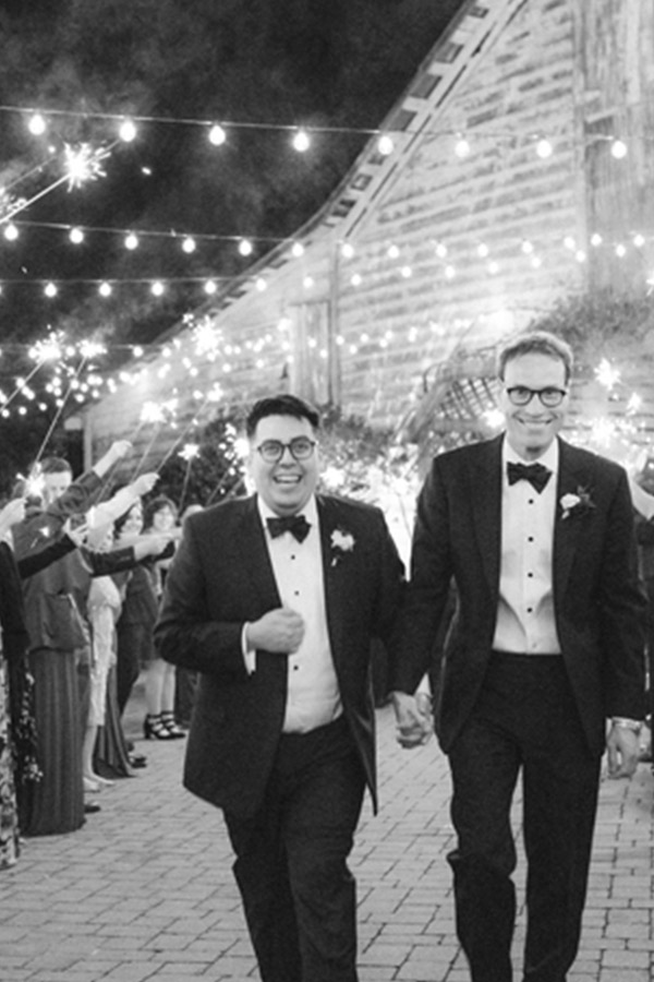 Two grooms in tuxedos, smiling and holding hands, walk down an outdoor path under string lights and sparklers held by guests. The atmosphere is festive and celebratory, with people clapping in the background near a rustic building. The image is in black and white. Fearrington Village