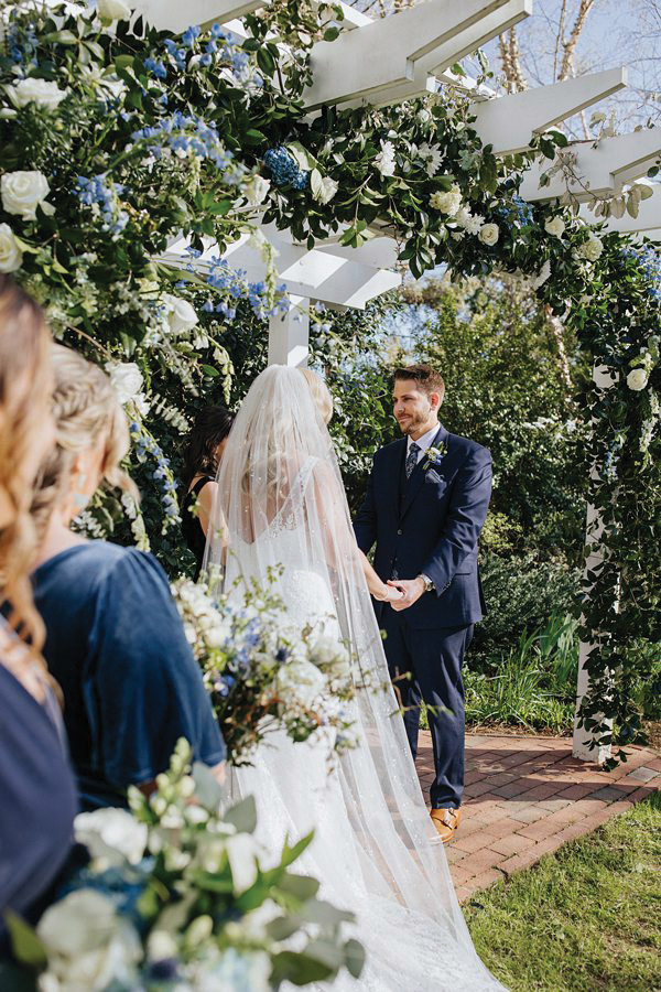 A bride and groom stand under a white arch adorned with green and blue floral decorations during their outdoor wedding ceremony. The bride, in a lace dress and veil, holds hands with the groom, who wears a navy suit. Bridesmaids carrying bouquets are partially visible. Fearrington Village