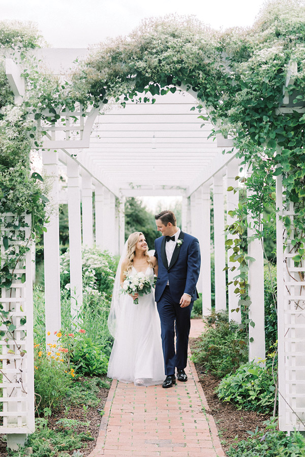 A bride and groom walk hand-in-hand down a brick path under a white pergola adorned with greenery and flowers. The bride wears a white dress and veil, holding a bouquet, while the groom is in a navy blue suit with a white boutonniere. Both smile happily. Fearrington Village