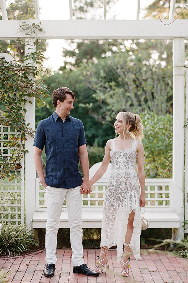 A couple stands hand in hand, smiling at each other under a white pergola in an outdoor garden. The woman is in a lacy white dress with strappy sandals, while the man wears a dark blue shirt and light-colored pants with black shoes. They appear happy and relaxed. Fearrington Village