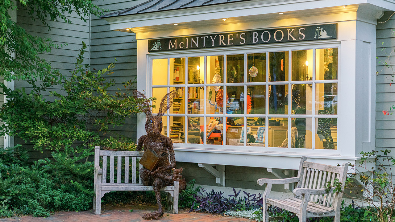 McIntyre's Books, a quaint bookstore, McIntyre's Books, featuring a large window displaying the interior. A unique tree-like sculpture holding an open book sits on a wooden bench in front, surrounded by green foliage and another empty wooden bench. Fearrington Village