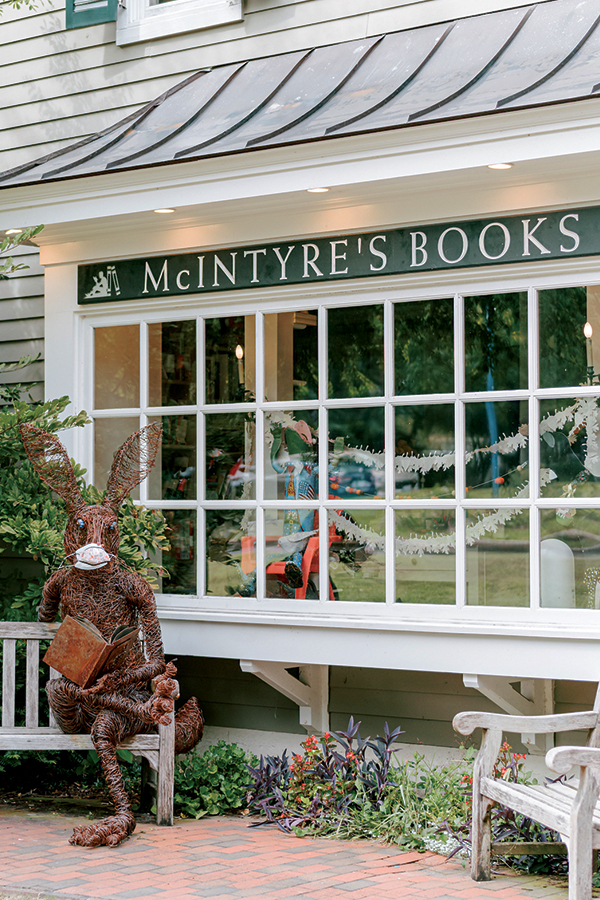 A bookstore named McIntyre's Books has a white-framed window with a sign above it. In front, there's a large sculpture of a rabbit made from twigs, sitting on a bench and holding an open book. Various plants adorn the area around the bench. Fearrington Village