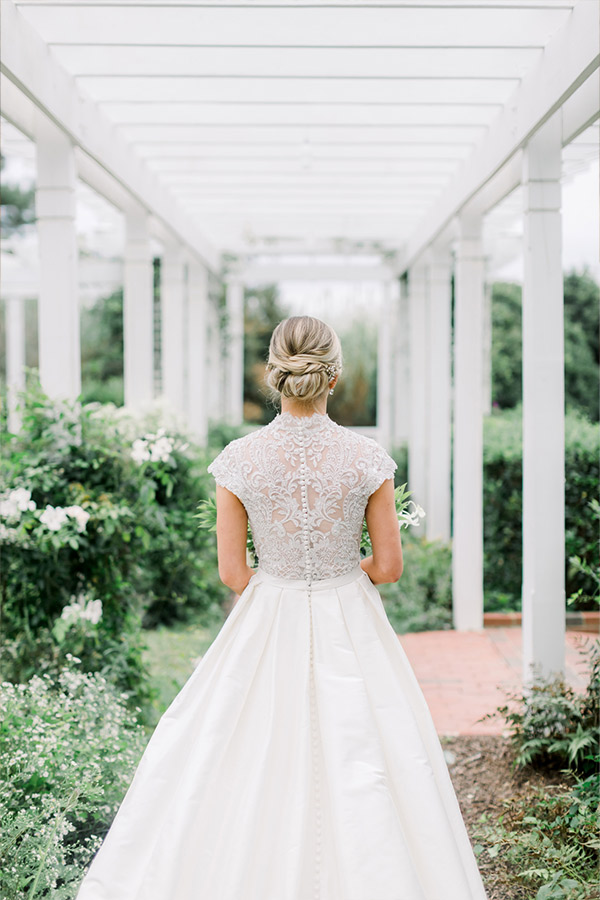 A bride stands with her back to the camera in an outdoor garden walkway framed by a white pergola. She wears an elegant dress with a lace back and holds a small bouquet. Her hair is styled in an intricate updo. White flowers and greenery surround the path. Fearrington Village