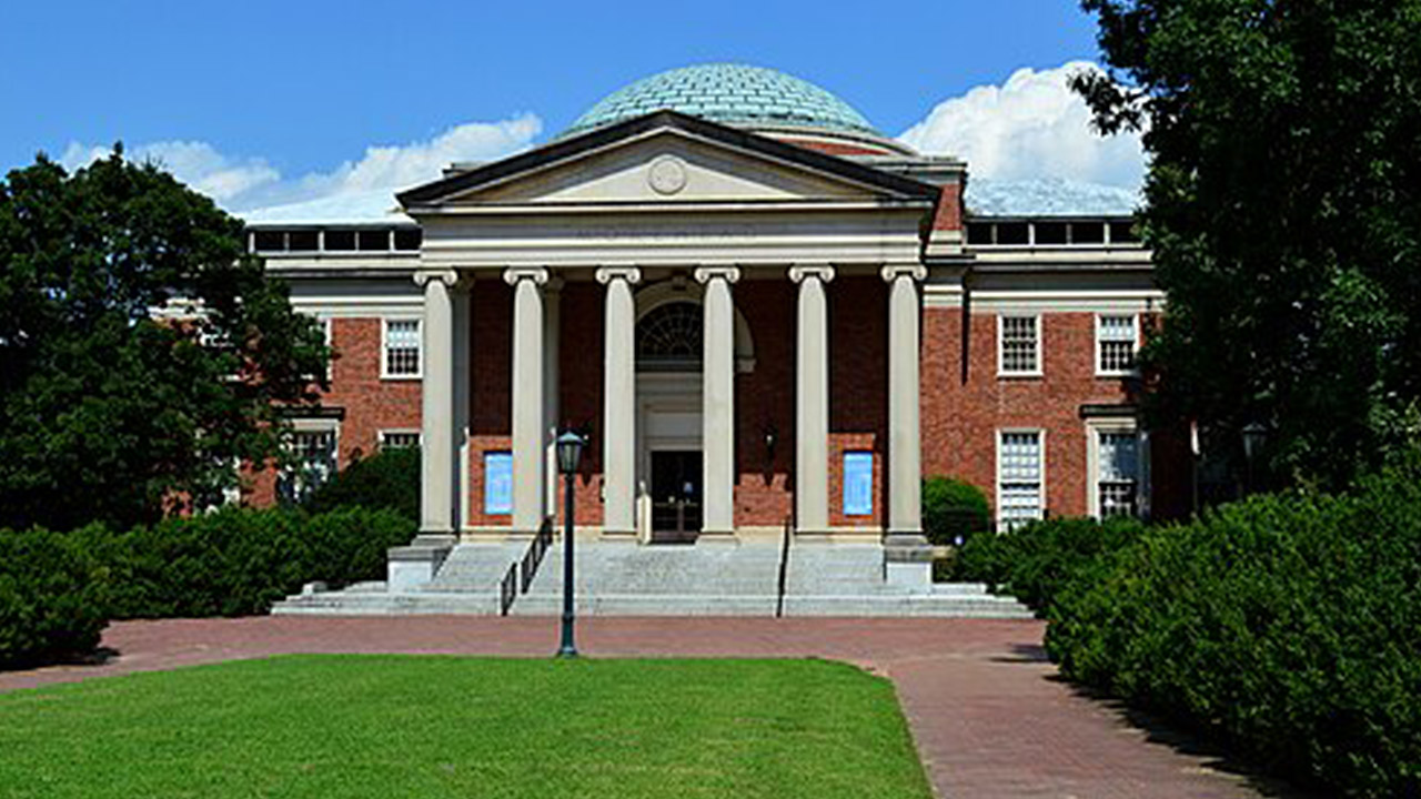 Morehead Planetarium. A stately red-brick building with a white portico featuring six columns supports a pediment, and a large dome is visible on the roof. The entrance is flanked by greenery and a lamppost stands in front, with a paved walkway leading up to the stairs. Fearrington Village