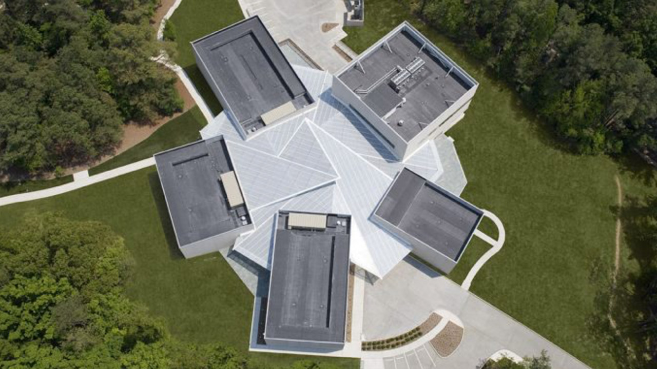 An aerial view of a modern, geometric building featuring a central structure with five protruding rectangular wings. The building is surrounded by green lawns and trees, with paved paths and parking areas visible. Fearrington Village