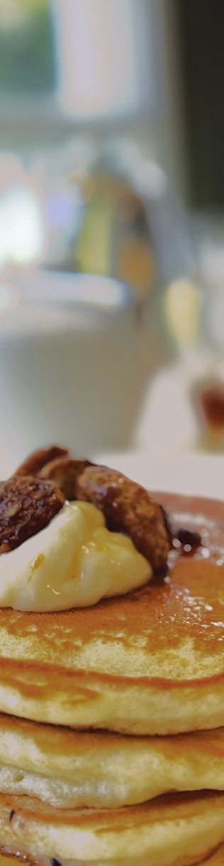 A close-up view of a stack of fluffy pancakes topped with a dollop of whipped cream and pieces of nuts. In the blurred background, there's a white cup and a jar, suggesting a cozy breakfast setting. Fearrington Village