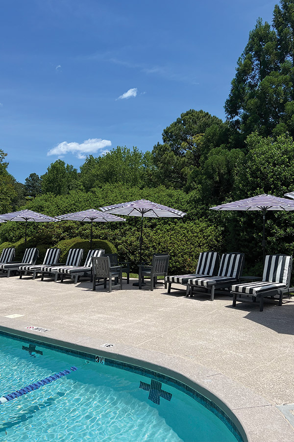 A poolside area featuring several black and white striped lounge chairs under matching umbrellas. The pool water is clear and blue, surrounded by a hedge of green trees and bushes. The sky is bright and clear with a few clouds. Fearrington Village