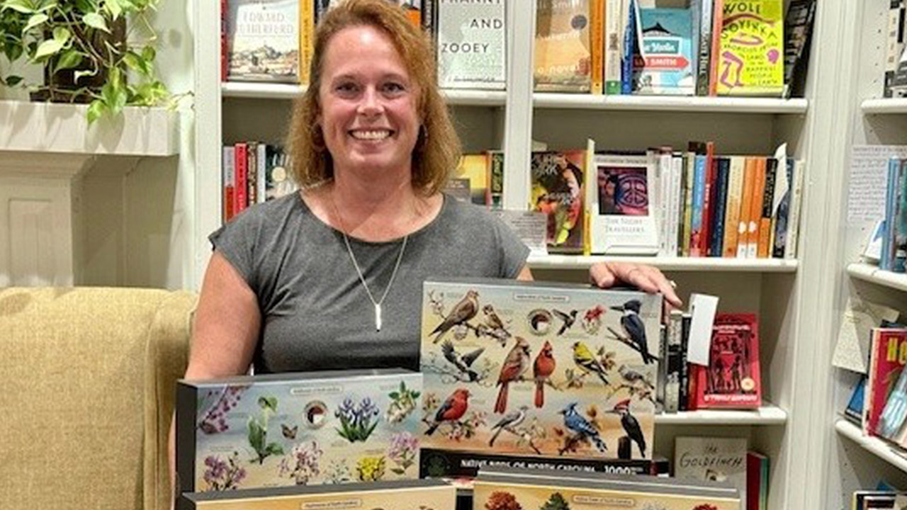 A woman with shoulder-length red hair stands in front of bookshelves filled with various books. She is smiling and holding up five bird-themed puzzles. The background includes more books and plants on a shelf. Fearrington Village