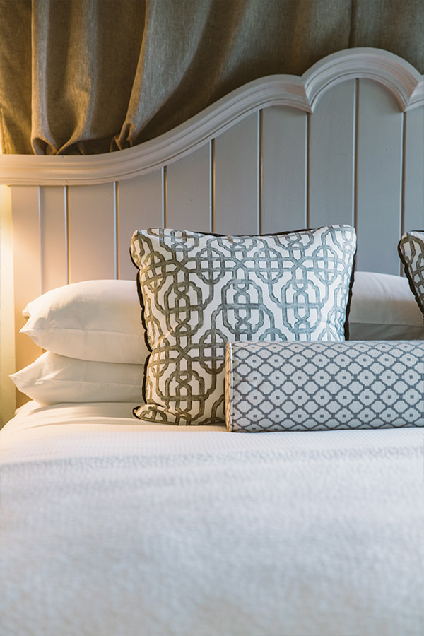 A neatly made bed with a white headboard. The bed features decorative pillows: two square pillows with a geometric pattern in white and gray, and a rectangular bolster pillow with a smaller, similar pattern. The headboard has a wavy top edge, and there's soft lighting. Fearrington Village