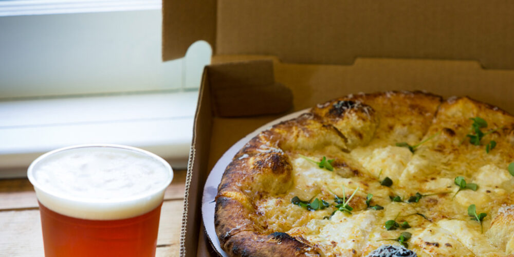 A freshly baked pizza with herbs and melted cheese sits in an open cardboard pizza box placed on a wooden table. Next to the box, there is a pint of beer with a frothy head, positioned close to a window with natural daylight coming through. Fearrington Village