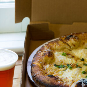 A freshly baked pizza with herbs and melted cheese sits in an open cardboard pizza box placed on a wooden table. Next to the box, there is a pint of beer with a frothy head, positioned close to a window with natural daylight coming through. Fearrington Village