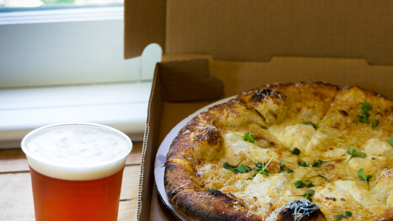 A freshly baked pizza with herbs and melted cheese sits in an open cardboard pizza box placed on a wooden table. Next to the box, there is a pint of beer with a frothy head, positioned close to a window with natural daylight coming through. Fearrington Village