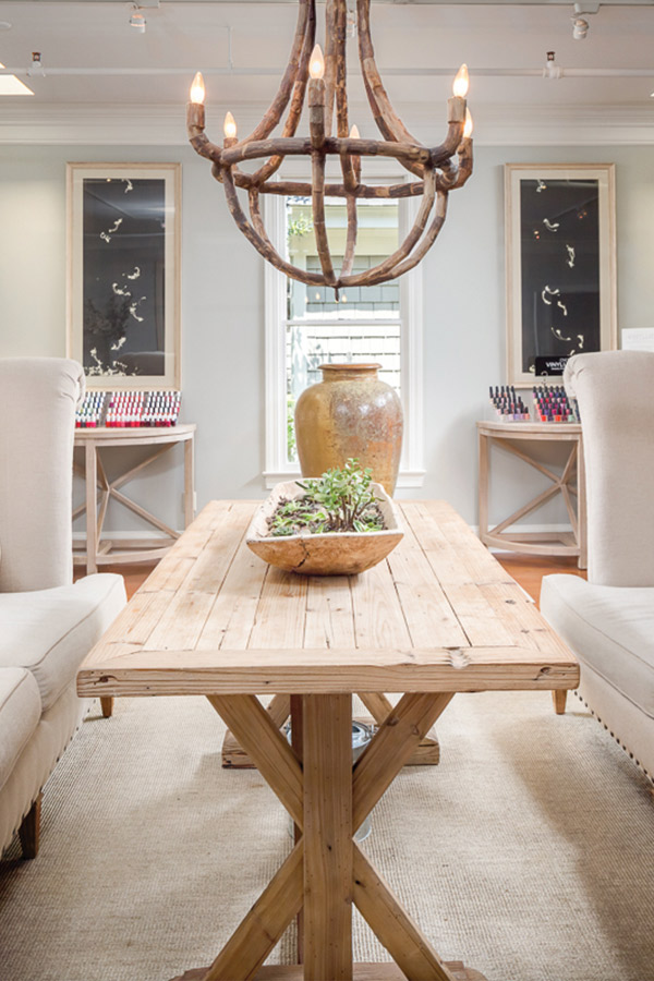 A rustic dining room features a wooden table with a succulent centerpiece, flanked by two cushy beige chairs. A large wooden chandelier hangs overhead. Shelves and a window are seen in the background. The room has a cozy and inviting atmosphere. Fearrington Village