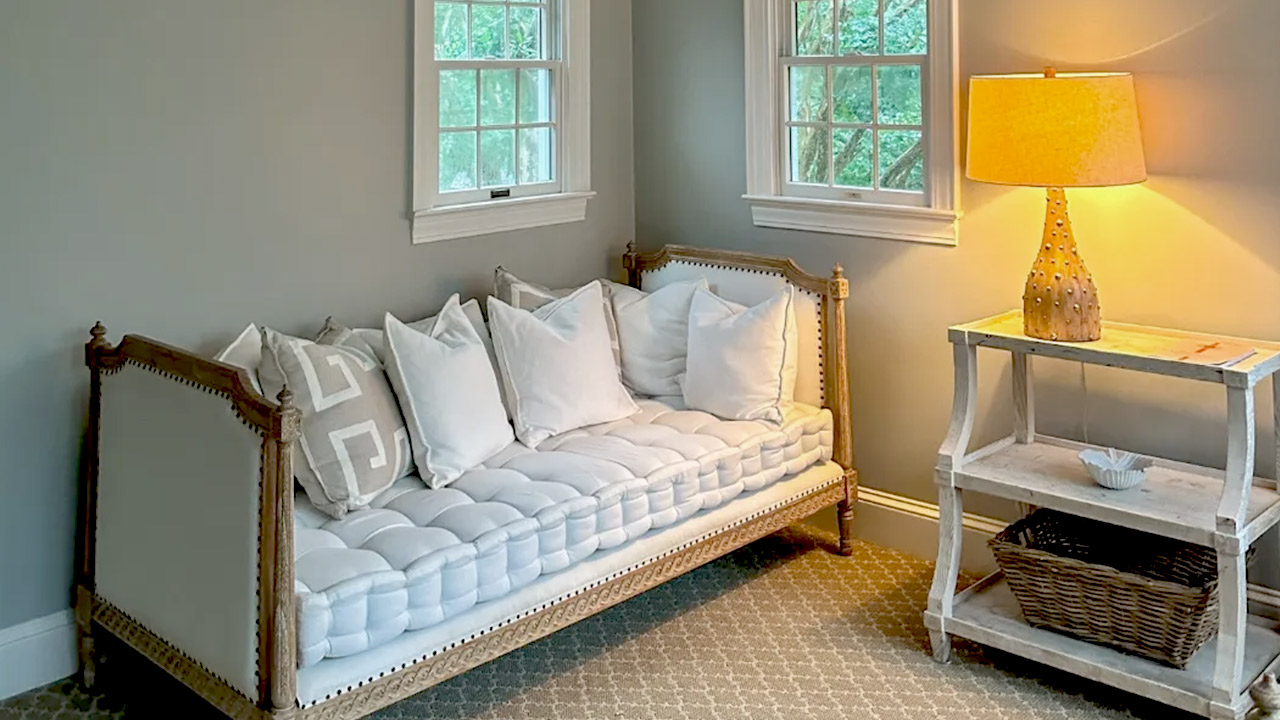 The Spa at Fearrington. A cozy room with a cushioned daybed adorned with multiple white pillows, placed under two double-hung windows. A wooden side table with a rustic lamp and a wicker basket on the lower shelf is positioned to the right of the daybed. Fearrington Village