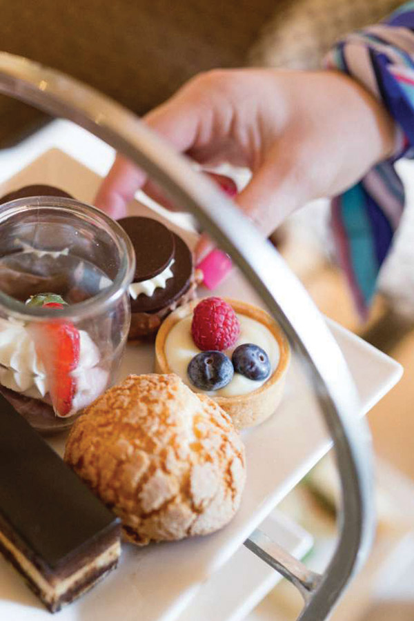 A close-up of a dessert tray featuring various pastries. The tray includes a chocolate layered dessert, a cream puff, a fruit tart with raspberries and blueberries, and a small glass container filled with whipped cream and a strawberry. A hand is reaching towards a cookie sandwich with cream. Fearrington Village