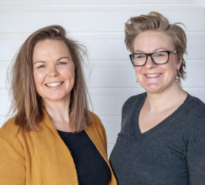 Tenille & Kim Chalmers. Two smiling individuals stand side by side in front of a white wooden background. The person on the left has long, straight hair and wears a mustard-colored cardigan over a black shirt. The person on the right has short hair, glasses, and wears a dark grey v-neck top. Fearrington Village