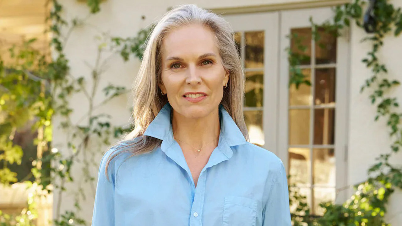 A woman with long gray hair wearing a light blue button-up shirt stands outdoors in front of a building with glass-paneled doors and greenery. She is smiling gently at the camera. Fearrington Village