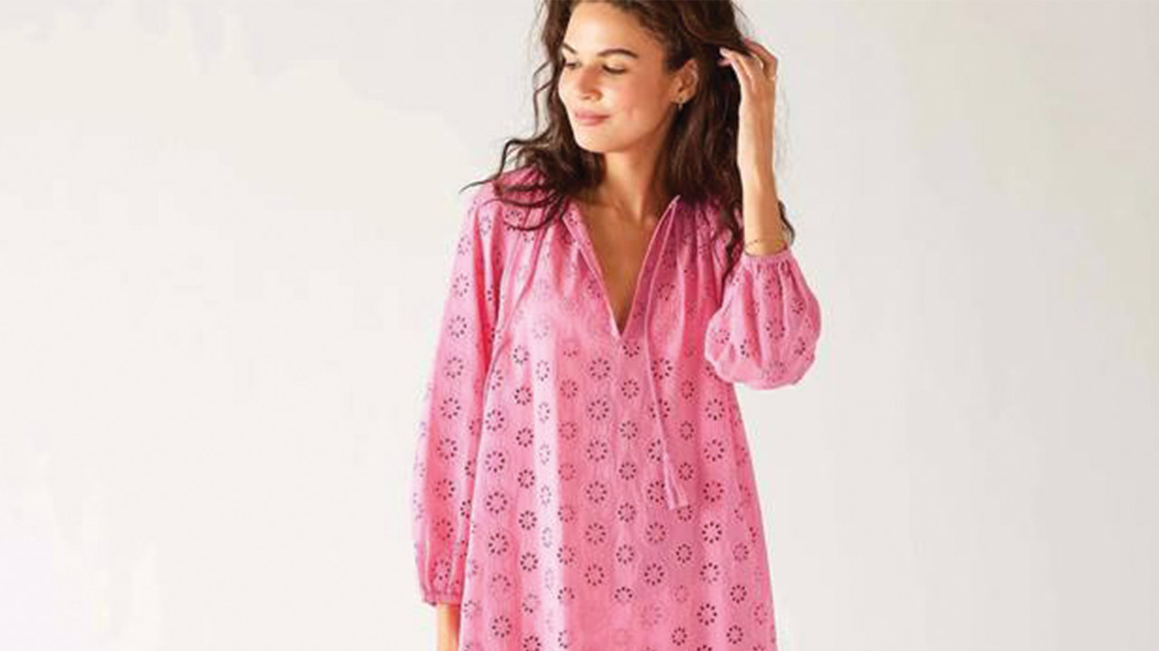 A woman stands against a plain background, looking to the side with a gentle smile. She is wearing a pink, long-sleeved dress with a floral eyelet pattern and a V-neckline. Her wavy hair is down, and she has one hand raised to tuck her hair behind her ear. Fearrington Village