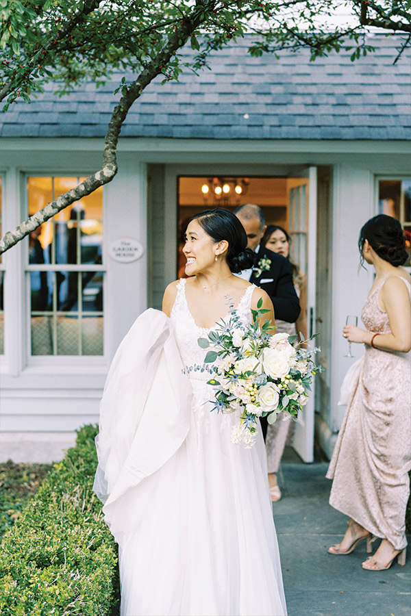 A bride in a white gown stands outside a building, smiling and holding a bouquet of white roses and greenery. Behind her, guests in formal attire gather near the entrance. She is lifting part of her dress with one hand. The scene appears joyful and celebratory. Fearrington Village