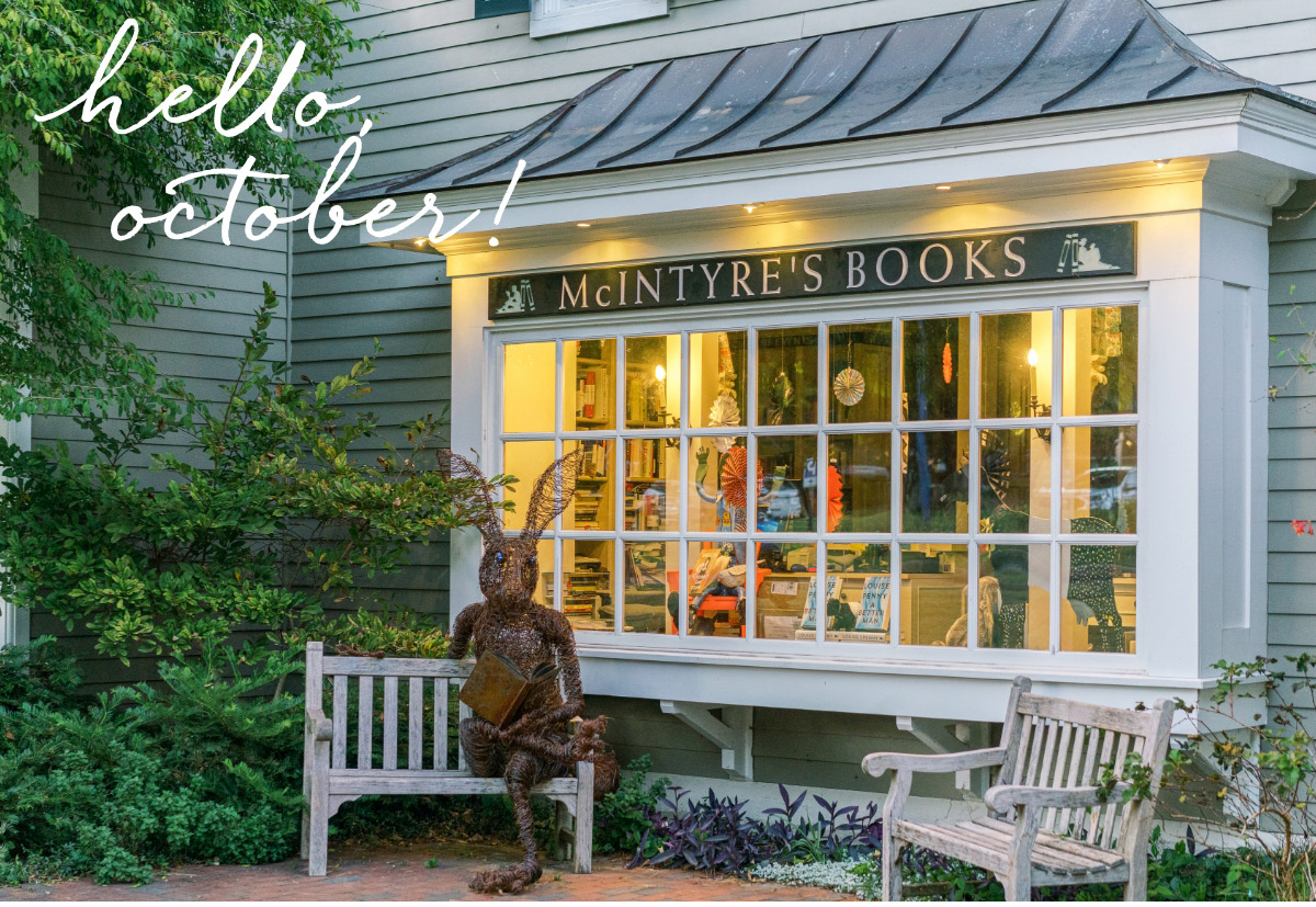 A shop window labeled "McIntyre's Books" is seen with warm interior lights. Outside, a bench and a sculpture made of twigs are displayed. The scene is lush with greenery, and the handwritten text "hello october!" is overlaid at the top left. Fearrington Village