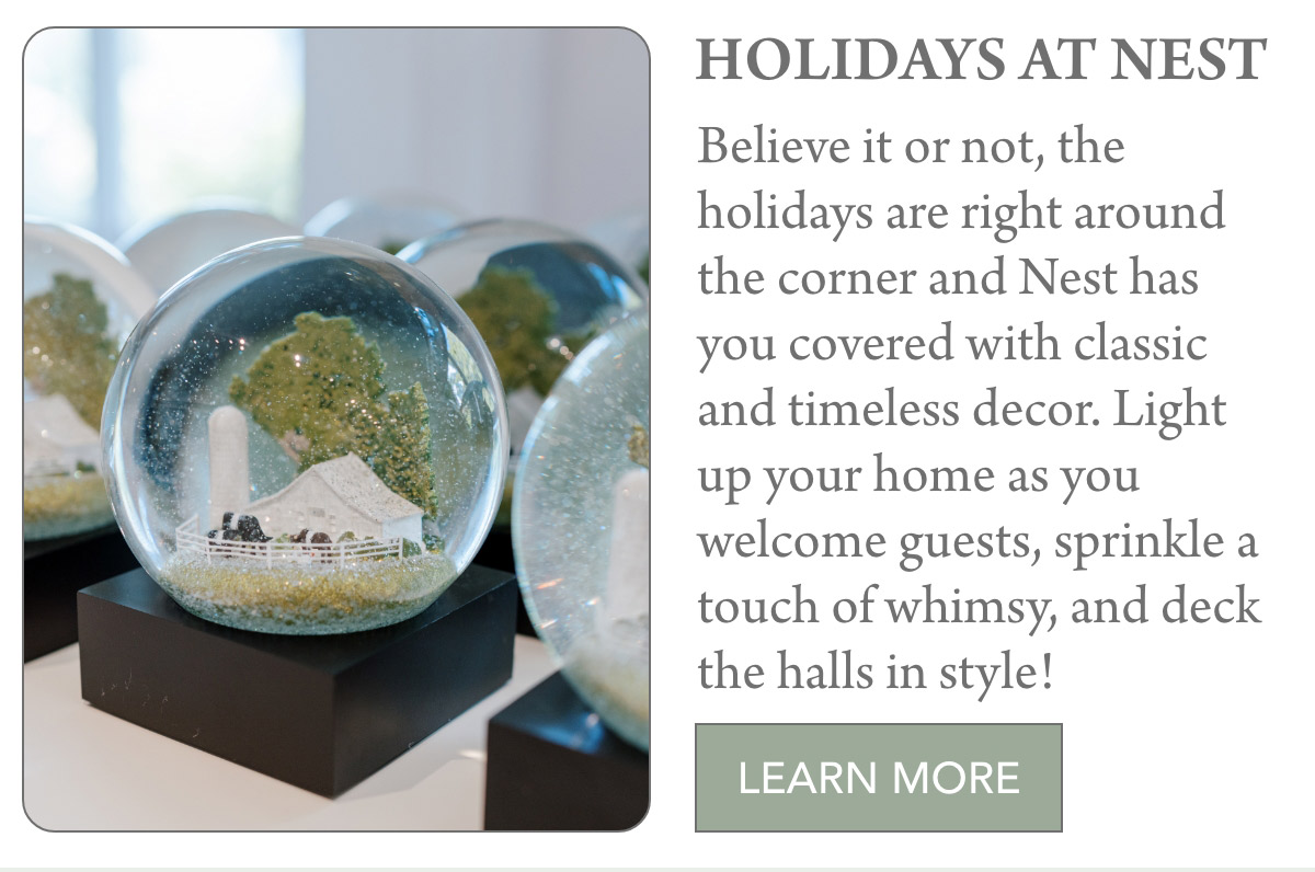 Several decorative snow globes on black bases are displayed in the foreground. The text on the right promotes holiday decor from Nest, suggesting their products add classic and timeless style. A green "Learn More" button is at the bottom of the text block. Fearrington Village