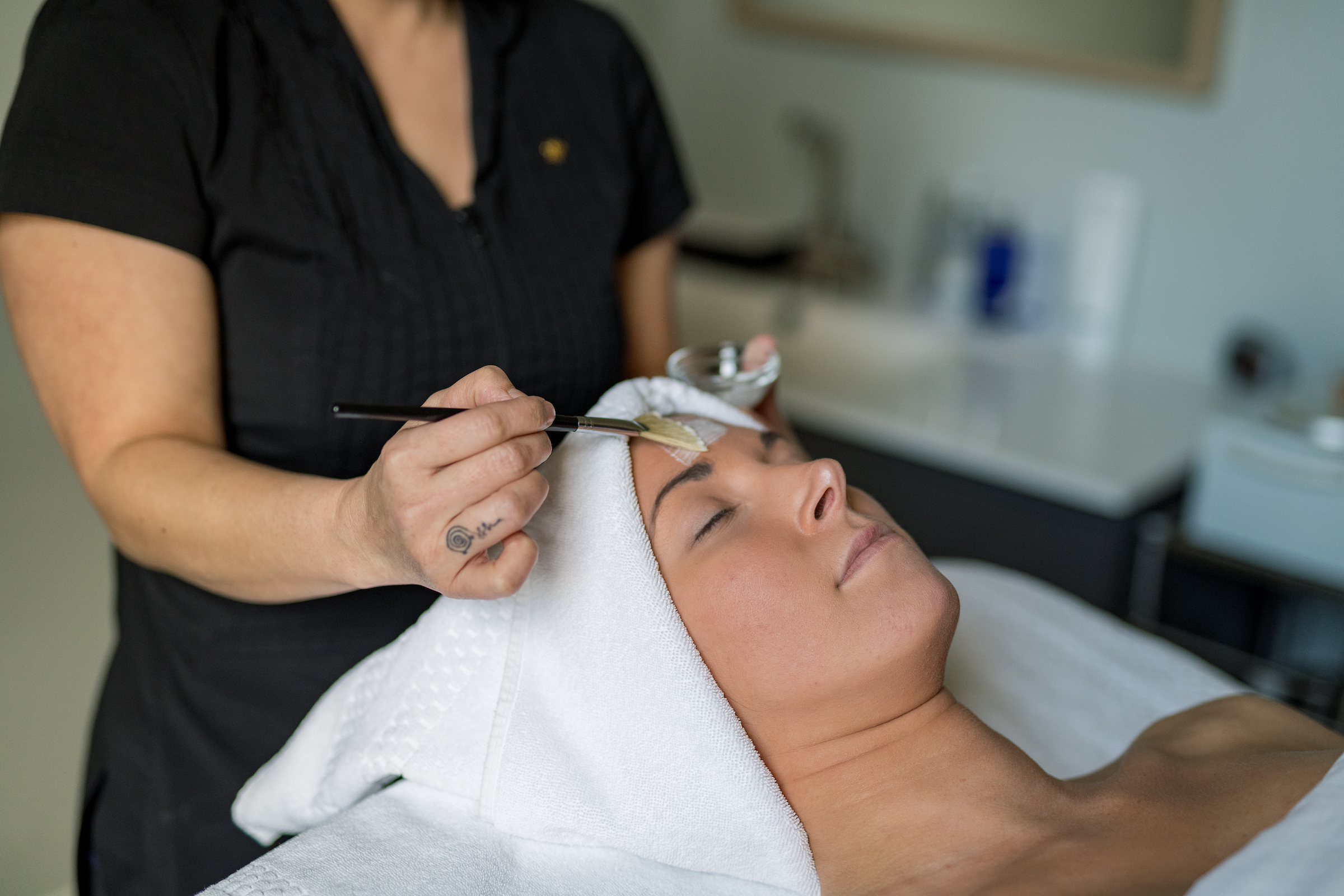 A person with a towel wrapped around their head lies on a spa table, receiving a facial treatment. Another person applies a face mask with a brush. The setting is a clean and modern spa or treatment room. Fearrington Village