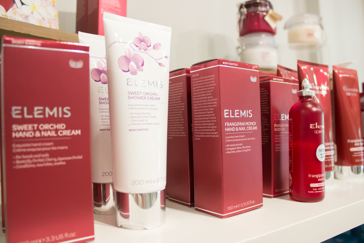 A display of ELEMIS skincare products, including Sweet Orchid Hand & Nail Cream, Exotic Shower Cream, and Frangipani Monoi Hand & Nail Cream. The products are in red and white packaging and are neatly arranged on a shelf. Fearrington Village