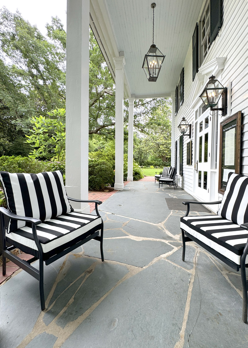 A covered porch with white columns, black and white striped cushioned chairs, and lantern-style lights hanging from the ceiling. The ground is paved with large, uneven stone tiles. Lush greenery surrounds the porch area. The house exterior is white with dark shutters. Fearrington Village