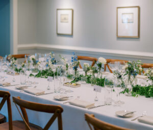 A long dining table is elegantly set with white tablecloth, white napkins, and crystal glassware. Greenery and white floral arrangements run down the center. Wooden chairs surround the table. Framed pictures hang on the light grey walls in the background. Fearrington Village