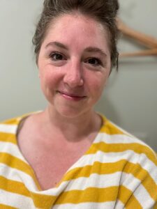 A person with light skin and brown hair is smiling at the camera. They are wearing a yellow and white horizontally striped shirt. The background is blurred, with a hanging rack visible on the wall. Fearrington Village