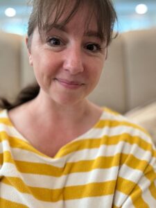 A woman with brown hair and bangs smiles at the camera. She is wearing a yellow and white striped top. The background is softly blurred, suggesting an indoor setting. Fearrington Village