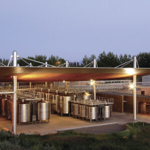 An outdoor wine production facility with large metal fermentation tanks under a modern canopy roof, surrounded by greenery, possibly vineyards, at dusk. The area is lit by artificial lights and features a combination of industrial and natural elements. Fearrington Village