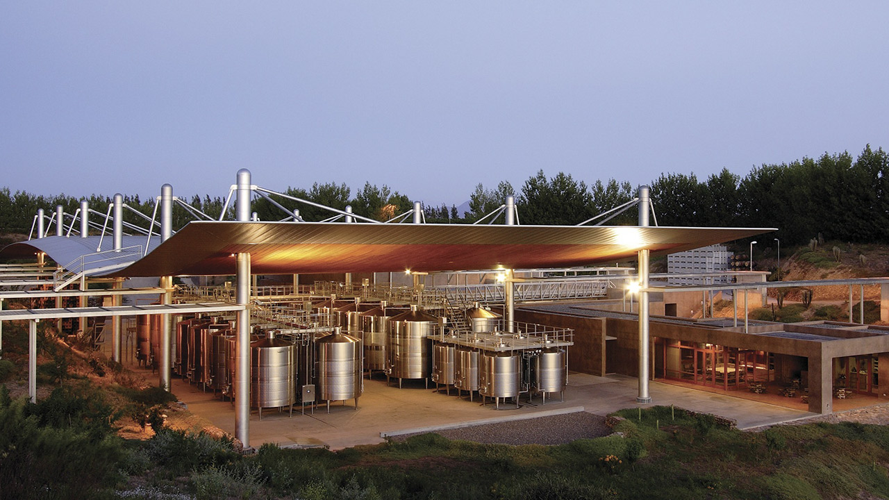 An outdoor wine production facility with large metal fermentation tanks under a modern canopy roof, surrounded by greenery, possibly vineyards, at dusk. The area is lit by artificial lights and features a combination of industrial and natural elements. Fearrington Village