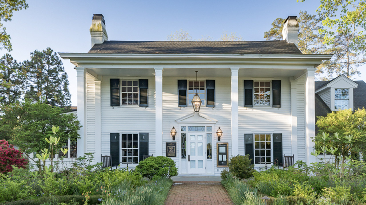 The Fearrington House Restaurant. A two-story white house with a gabled roof, white columns, and dark shutters. The front entrance features a lantern, surrounded by lush greenery and a red-brick walkway leading up to the door. Mature trees and shrubbery frame the property in the background. Fearrington Village