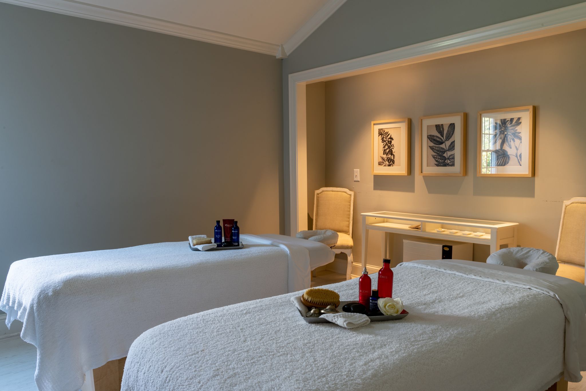 A serene spa room with two massage tables covered in white sheets. Each table has neatly arranged towels, oils, and brushes. The room features soft lighting, beige walls, framed botanical artwork, and two chairs near a table in the background. Fearrington Village