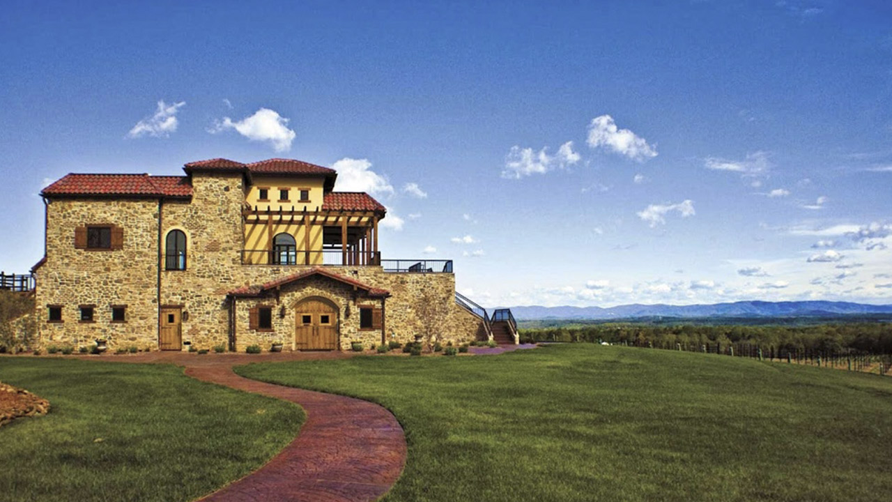 A large, rustic stone house with a red-tiled roof sits on a vast, manicured lawn. A curved brick pathway leads up to the house. In the background, there are rolling hills and a blue sky dotted with scattered clouds. Fearrington Village