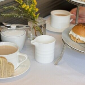 A cozy tea setup with two white cups of tea, one with biscuits shaped like hearts on the saucer. A small white pitcher, sugar bowl, and a tiered tray holding sandwiches are on the table. There is a small vase with yellow flowers in the background. Fearrington Village