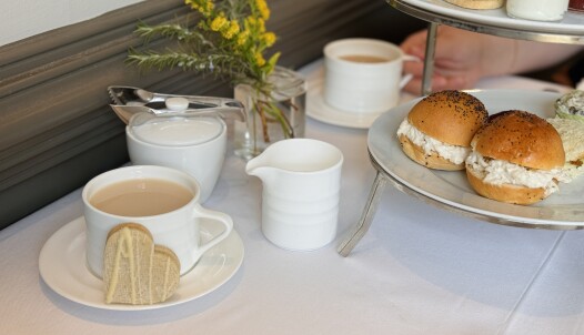 A cozy tea setup with two white cups of tea, one with biscuits shaped like hearts on the saucer. A small white pitcher, sugar bowl, and a tiered tray holding sandwiches are on the table. There is a small vase with yellow flowers in the background. Fearrington Village