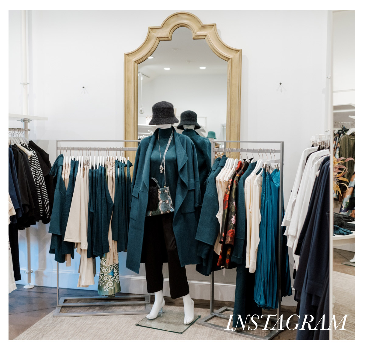 A clothing store display featuring a mannequin dressed in a teal coat, black hat, black pants, and white boots with a metallic handbag. Behind the mannequin, a large ornate mirror is visible, reflecting part of the store's interior. The word "INSTAGRAM" is written in the bottom right corner. Fearrington Village