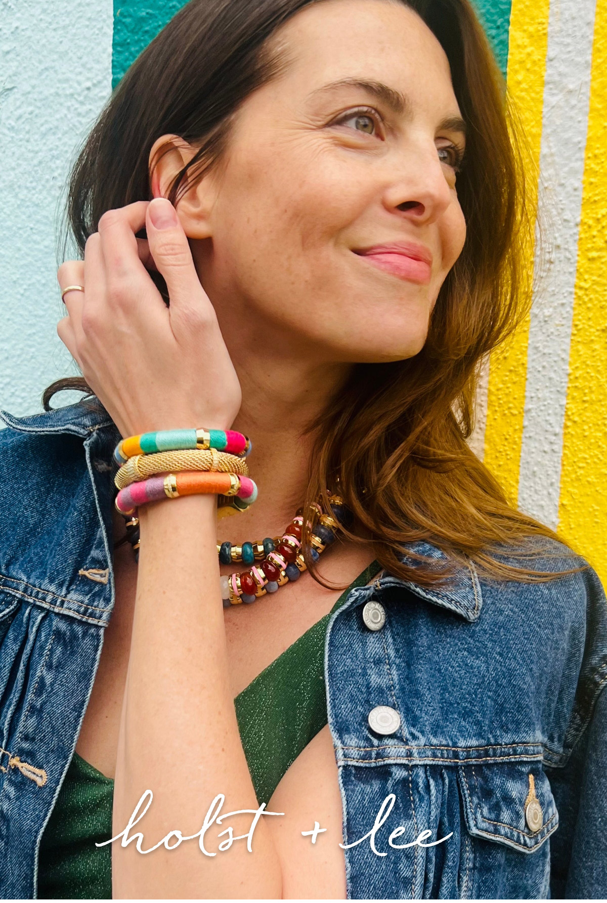 A woman with long brown hair, wearing a denim jacket over a green dress, has colorful bracelets and a necklace. She smiles while touching her ear, standing against a backdrop of pastel blue and yellow stripes. Text at the bottom reads "holst + lee. Fearrington Village