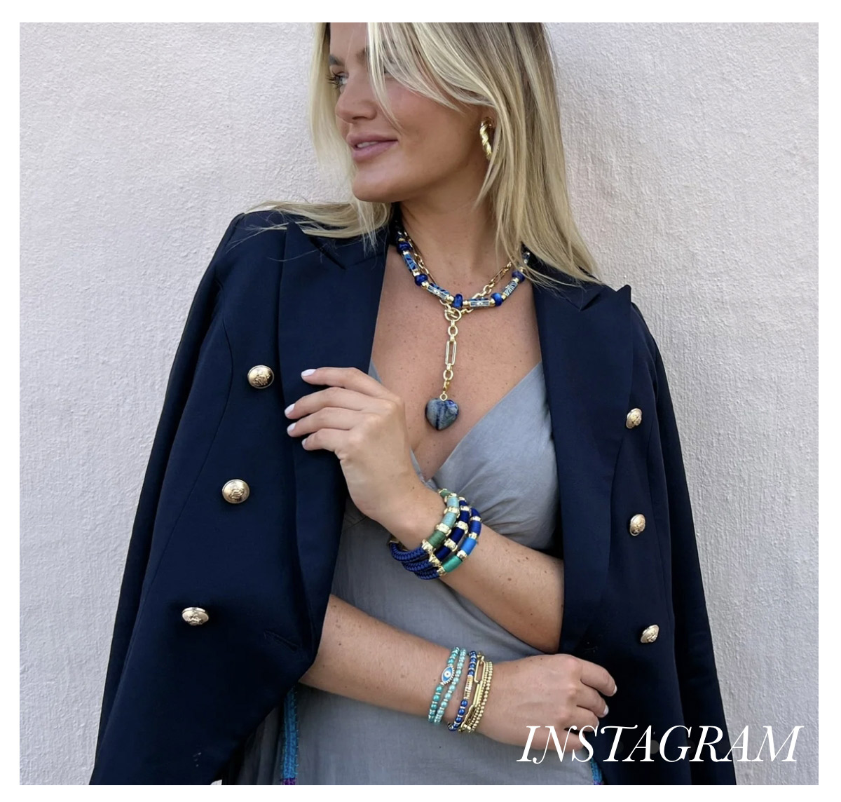 A blonde woman wearing a black jacket with gold buttons draped over her shoulders and a grey dress. She accessorizes with a multi-layered chain necklace, multiple bracelets, and hoop earrings. She stands against a plain light background, looking to the side. Fearrington Village