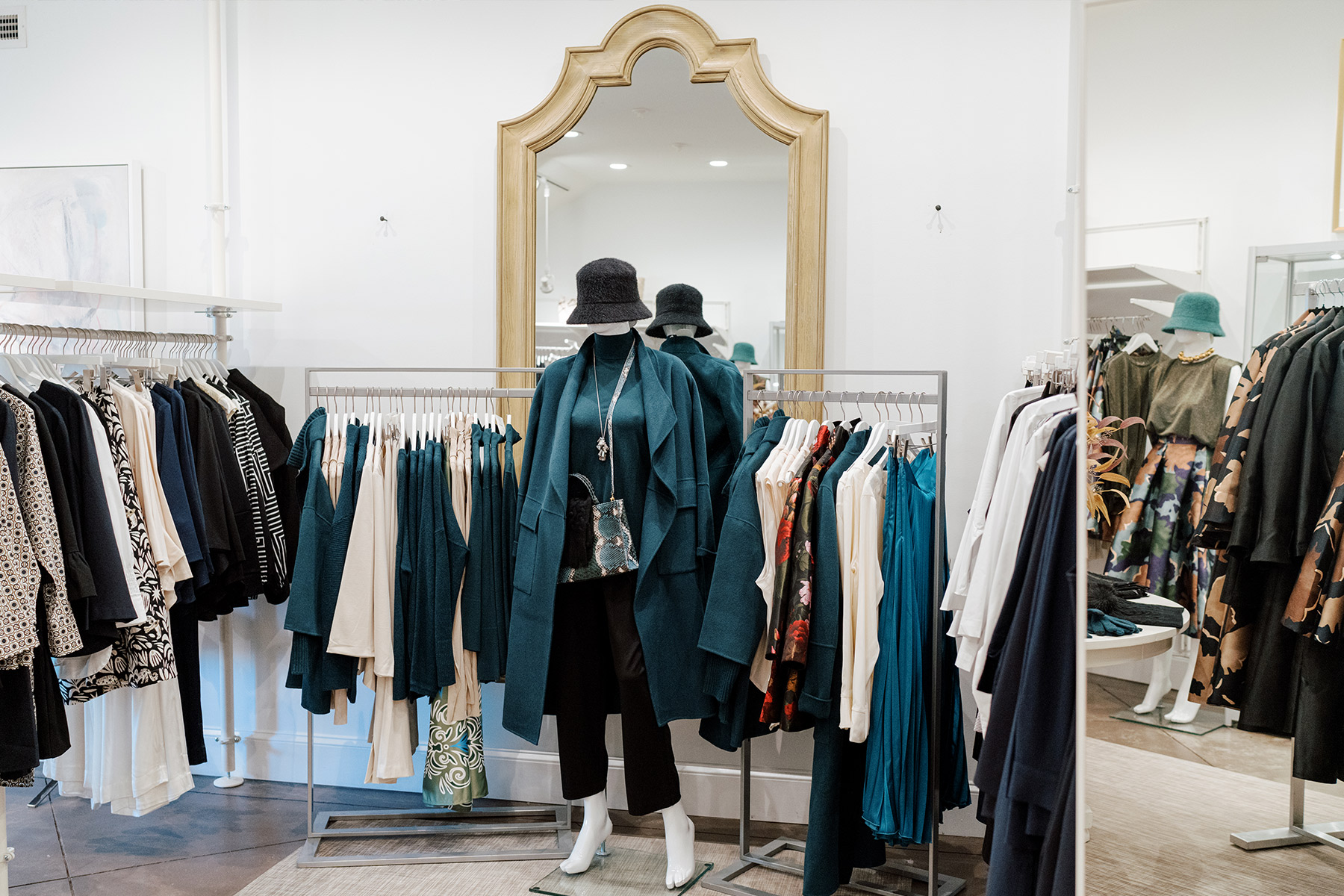 Dovecote. A chic clothing store display features a mannequin dressed in a teal coat, black hat, black pants, white boots, and a clear handbag. Surrounding racks showcase an array of clothes, including neutral and vibrant colors, with a large gold-framed mirror in the background. Fearrington Village