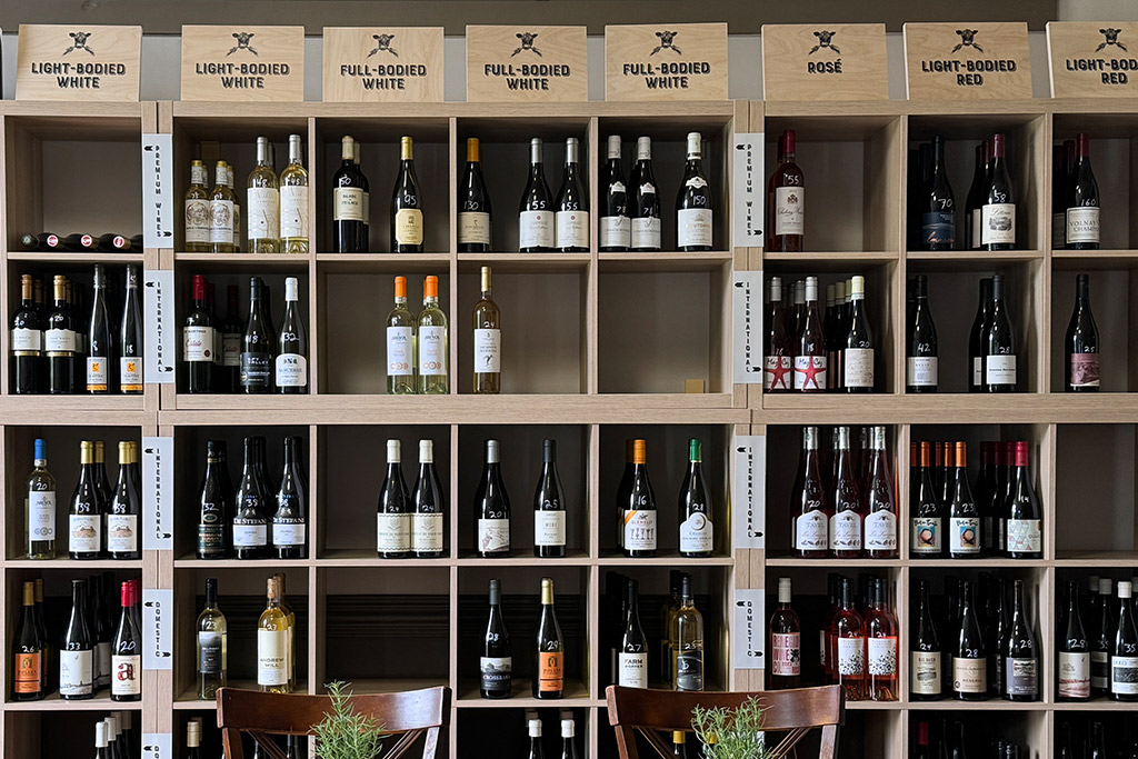 Galloway's Wine Shop. A neatly arranged shelf of wines in a store, categorized by type. The top row includes sections for light-bodied white, full-bodied white, rosé, and light-bodied red wines. Wine bottles fill each section, with empty chairs in front of the shelf. Fearrington Village