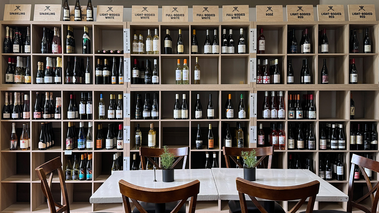 Galloway's Wine Shop. A neatly organized wine store display with wooden shelves filled with various wine bottles. The shelves are labeled by categories: Sparkling, Light-Bodied White, Full-Bodied Red, Rosé, Light-Bodied Red, and more. In the foreground are tables and chairs. Fearrington Village