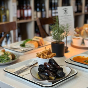 Galloway's Wine & Beer Bar. A table at a restaurant is set with various dishes including mussels, a bowl of fries, a creamy soup, sandwiches, and a salad with falafel. In the background, shelves filled with bottles of wine are visible. The table is decorated with a small potted plant. Fearrington Village