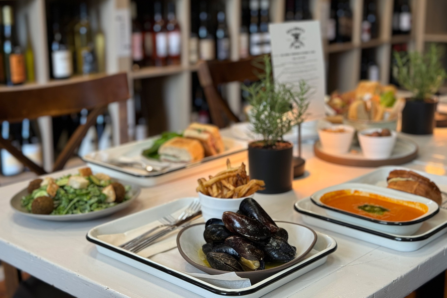 Galloway's Wine & Beer Bar. A table at a restaurant is set with various dishes including mussels, a bowl of fries, a creamy soup, sandwiches, and a salad with falafel. In the background, shelves filled with bottles of wine are visible. The table is decorated with a small potted plant. Fearrington Village