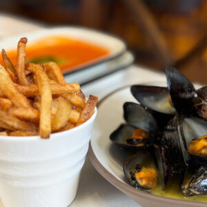 Galloway's Wine & Beer Bar. A plate of mussels in a bowl with sauce and a cup of French fries sit on a table. Another dish with tomato sauce can be seen in the background. The table is set with white and beige striped cloth. Fearrington Village