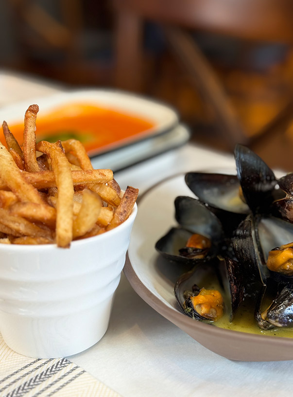 Galloway's Wine & Beer Bar. A plate of mussels in a bowl with sauce and a cup of French fries sit on a table. Another dish with tomato sauce can be seen in the background. The table is set with white and beige striped cloth. Fearrington Village