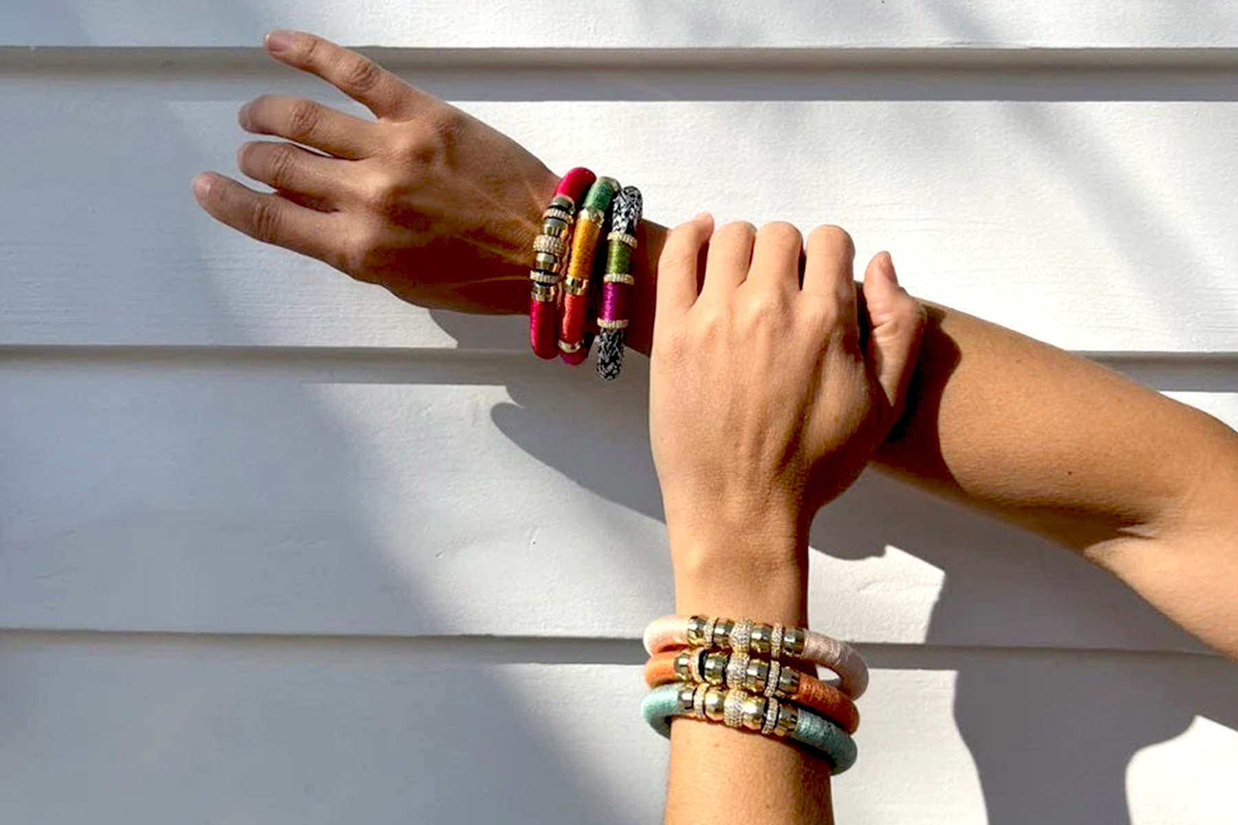 Two hands with colorful, stacked beaded bracelets are artistically positioned in front of a white, horizontally-paneled wall. The bracelets feature various colors and bead shapes, creating a vibrant and eclectic look. Shadows are cast on the wall, adding depth. Fearrington Village