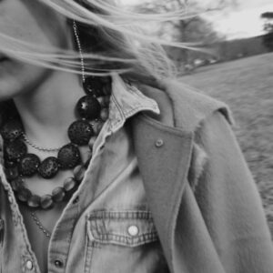 A black and white photo of a person wearing a denim jacket and layered necklaces, with long hair blowing in the wind. The background appears to be an open field with blurred trees and a cloudy sky. The image captures a sense of motion and casual style. Fearrington Village